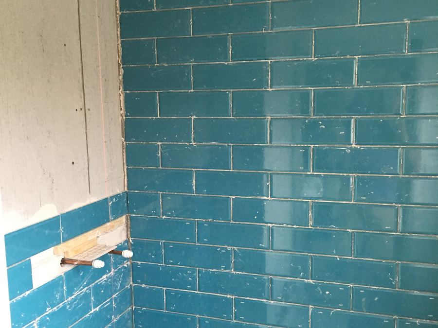 Teal brick-effect wall tiles being fixed in a bathroom, with a timber batten screwed across the wall to hold the first course level