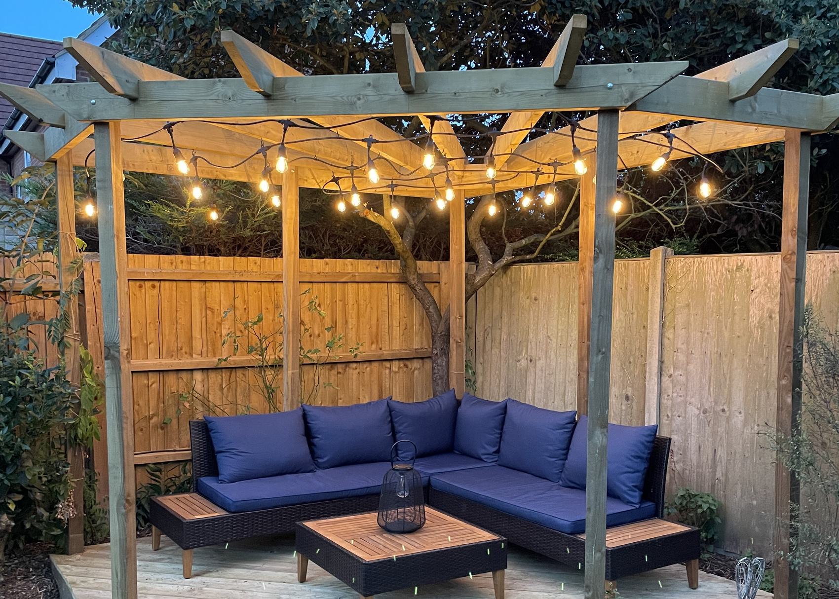 A timber pergola at dusk, its rafters cut to a fan tail at each end, festoon lights strung beneath, over a deck and corner sofa in a Cambridgeshire garden